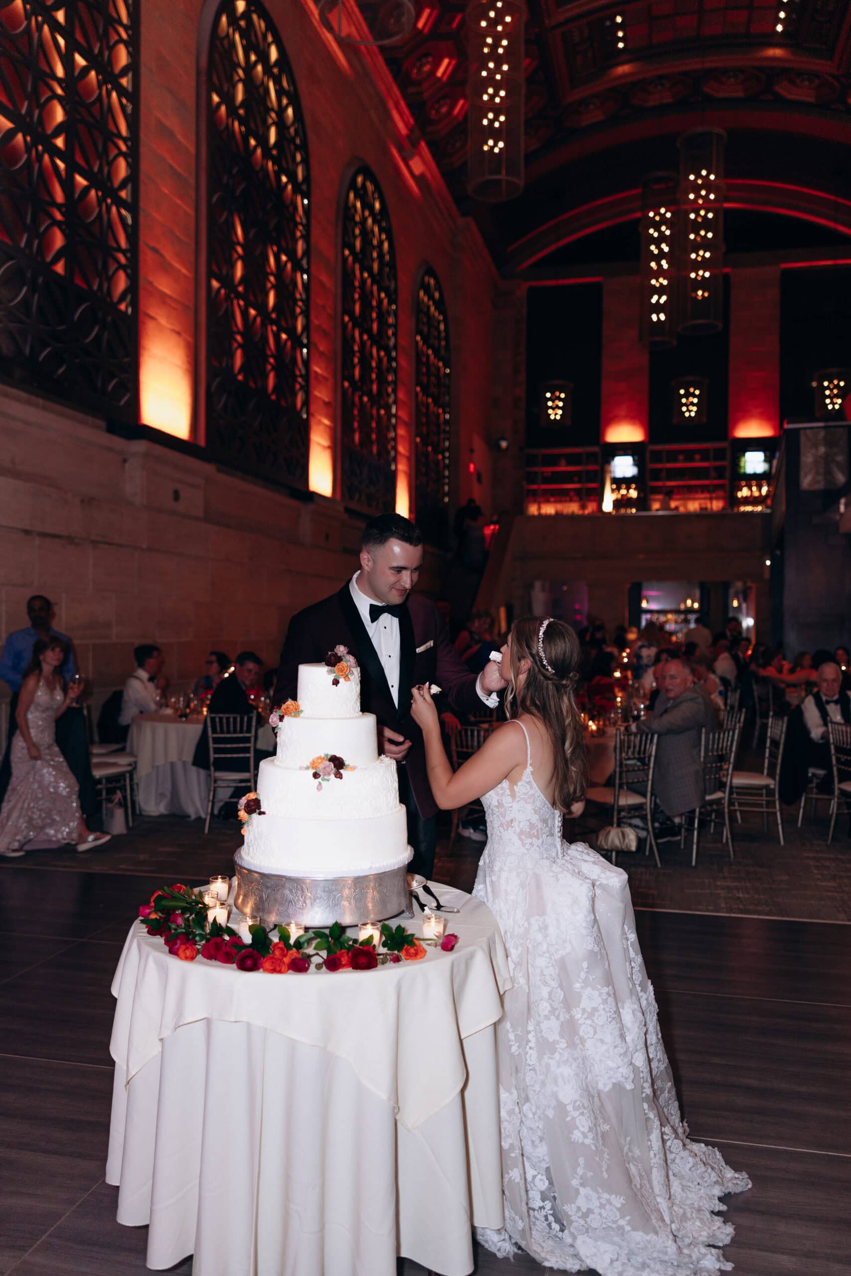 Bride in lace gown and groom in tuxedo feeding each other cake beside a four-tier white wedding cake at Union Trust, Philadelphia.