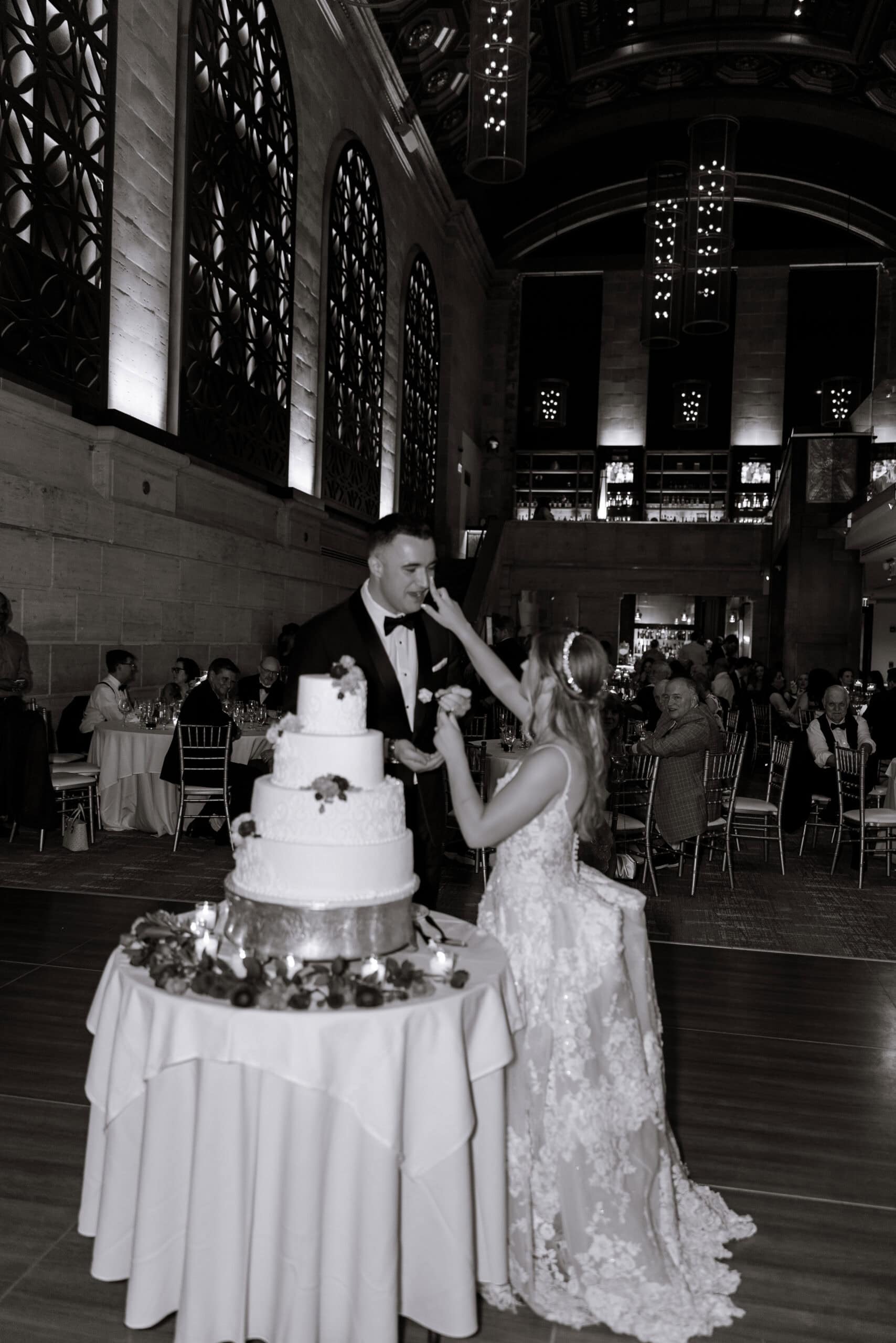 Bride in lace gown feeding groom cake during wedding reception at Union Trust in Philadelphia with ornate windows and guests seated in background.