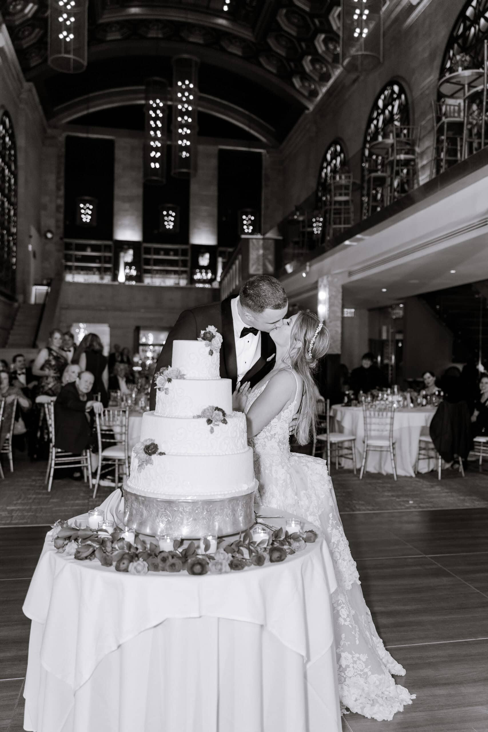 Black and white photo of a bride and groom kissing beside a four-tiered wedding cake adorned with flowers, in a grand hall with arched windows and chandeliers at Union Trust, Philadelphia.