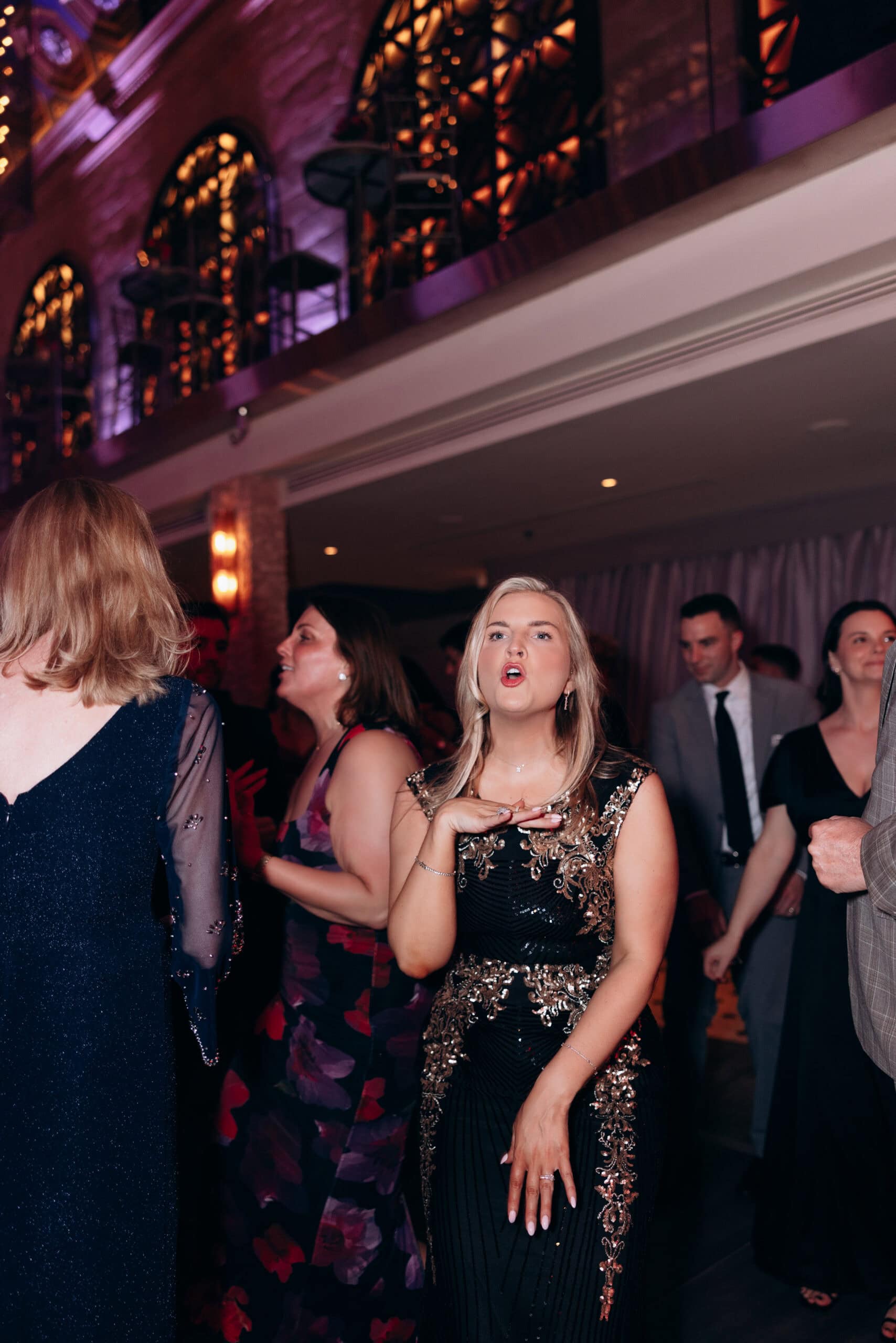 Woman in black and gold embroidered dress gesturing during an evening event at Union Trust, Philadelphia, with guests and ornate lighting in background.