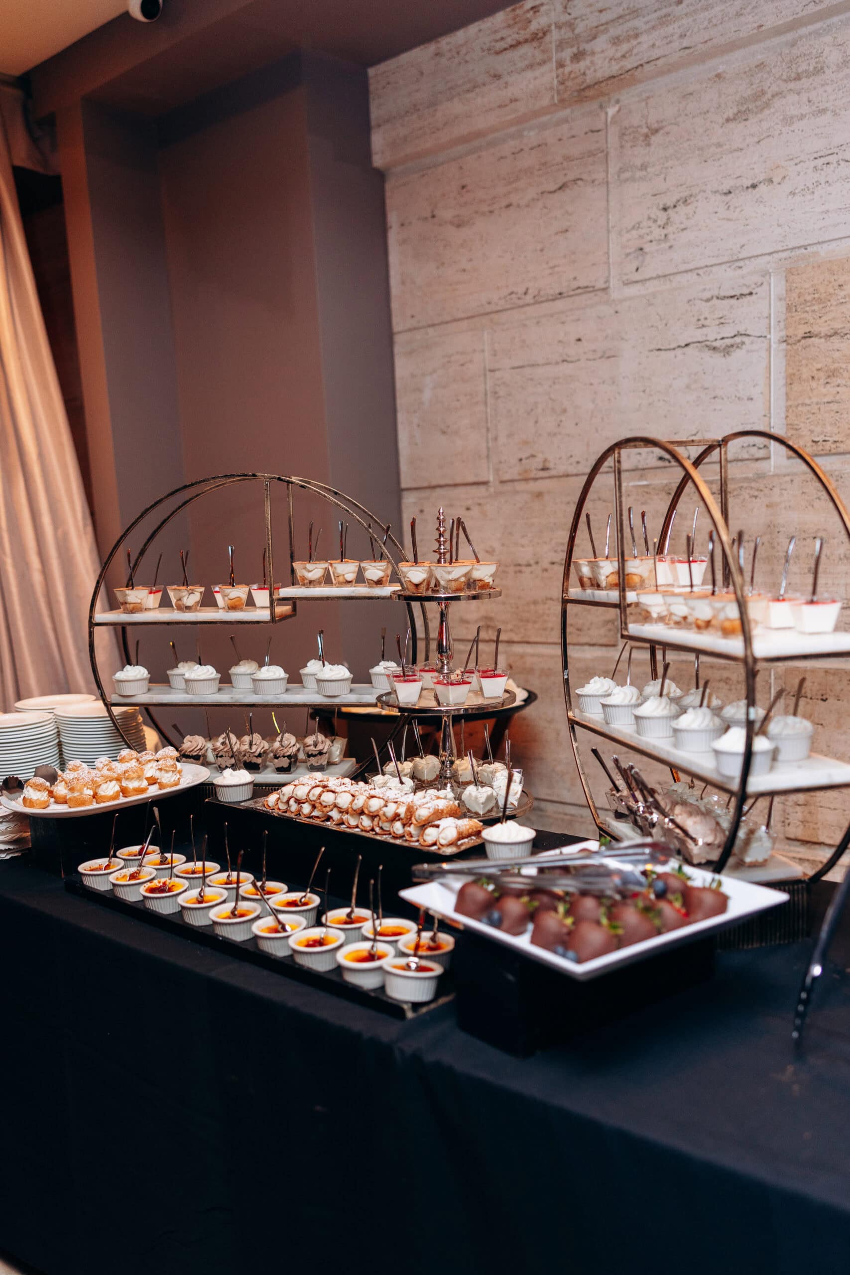 Tiered dessert display with assorted miniature pastries, creams, and chocolate-covered strawberries on black tablecloth against stone wall at Union Trust, Philadelphia.
