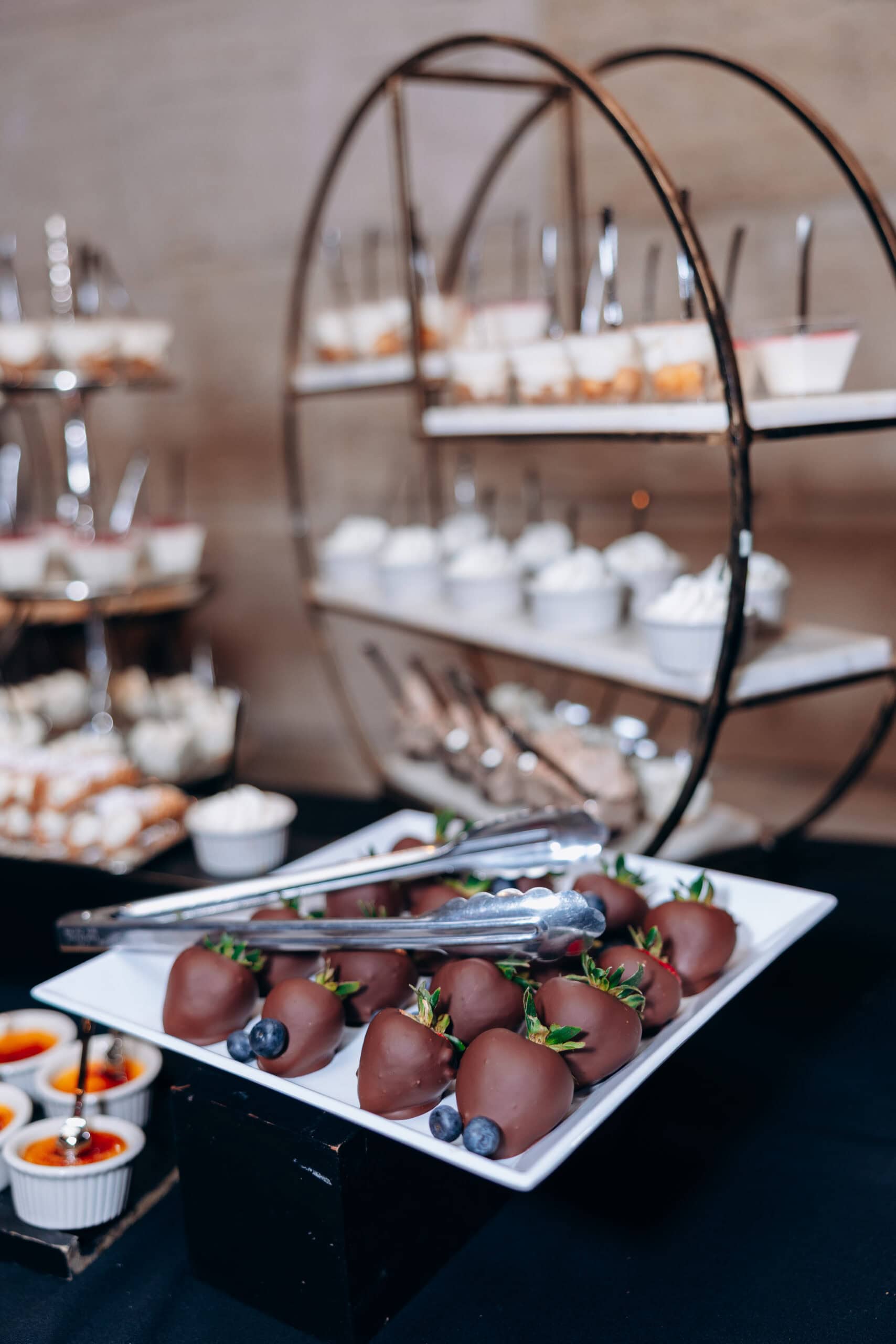 Close-up of a white rectangular platter with chocolate-covered strawberries and blueberries, with blurred tiered dessert trays in the background at a wedding venue.
