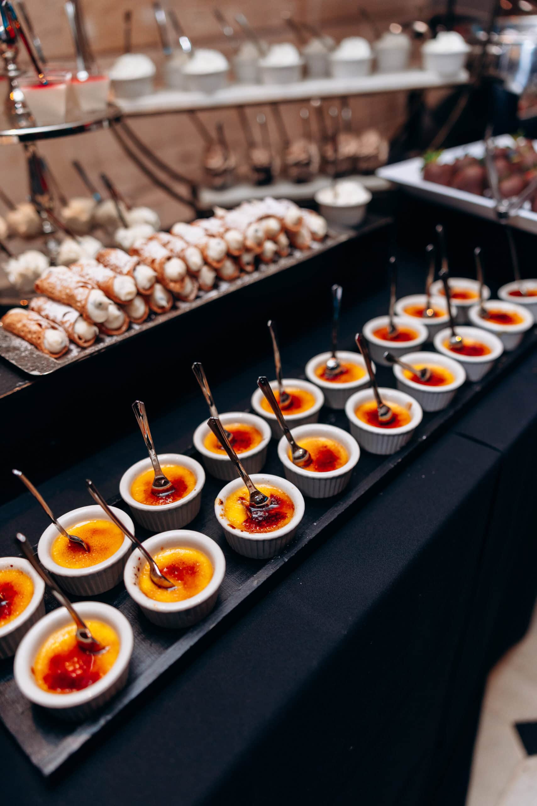 Rows of crème brûlée in white ramekins with spoons and powdered sugar-dusted cannoli on black trays at a wedding dessert table.