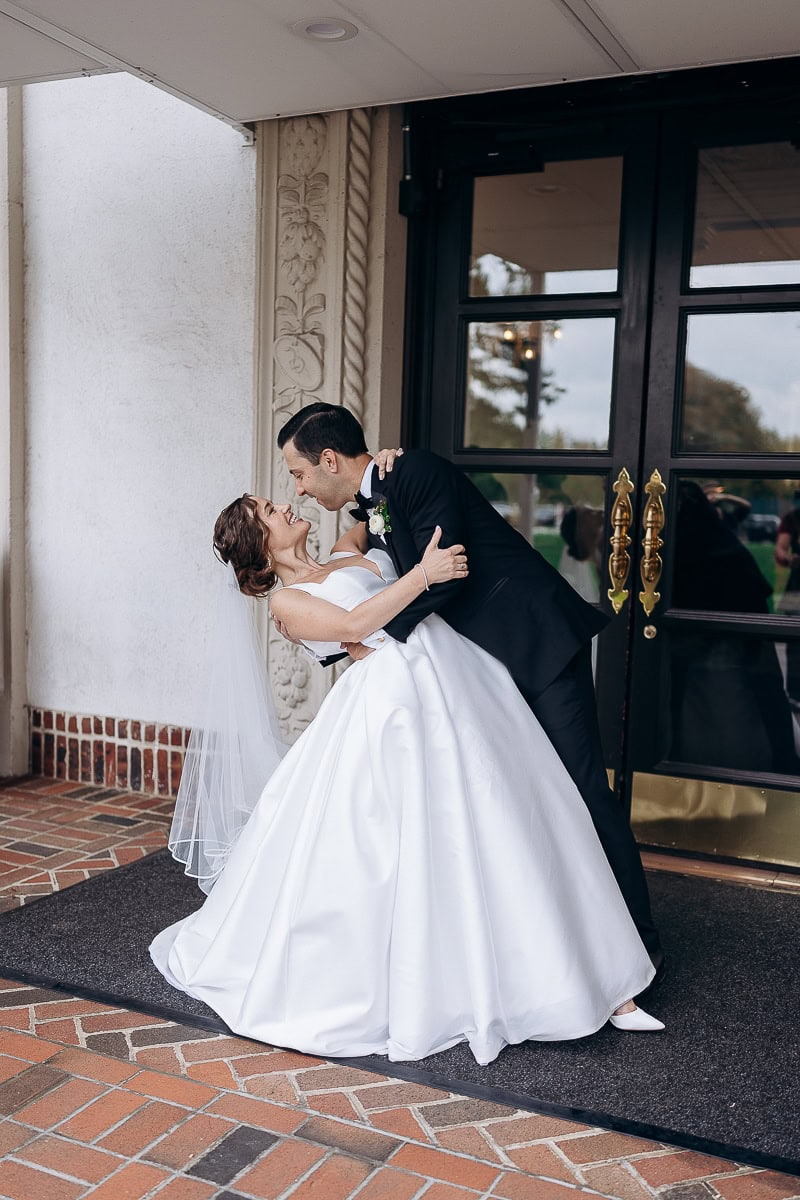 Bride in satin ballgown and veil dipped backward by groom in black tuxedo outside ornate entrance at Westchester Country Club, Rye, NY.