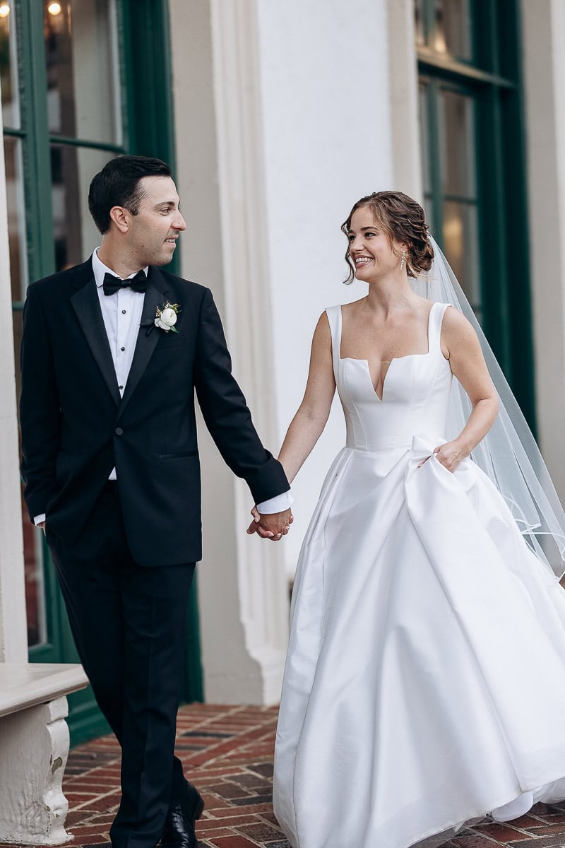 Bride in structured white gown with veil and groom in black tuxedo holding hands, walking outside Westchester Country Club, Rye NY.