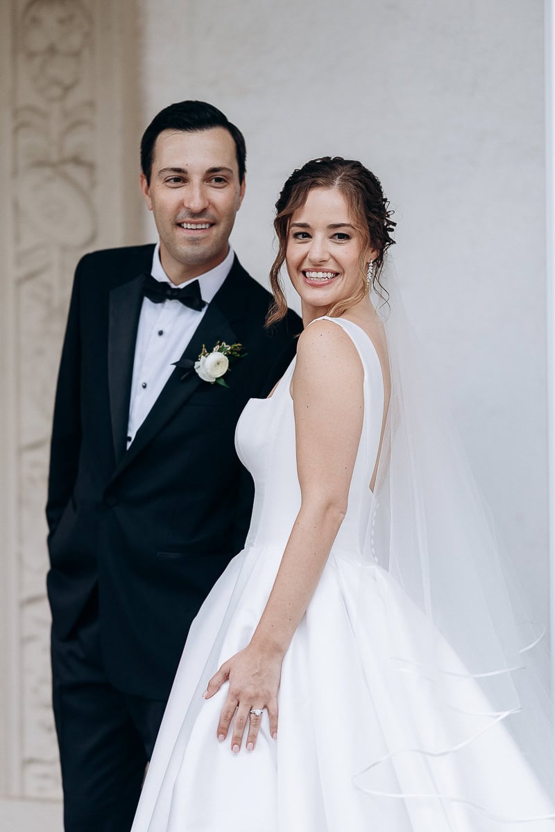 Bride in structured satin wedding dress with veil and groom in black tuxedo smiling, standing by ornate wall at Westchester Country Club, Rye, NY.