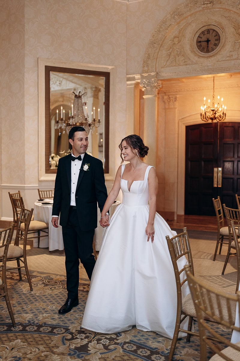 Bride in structured white satin gown and groom in black tuxedo holding hands, walking through an ornate room with classical architecture and chandeliers at Westchester Country Club, Rye, NY.
