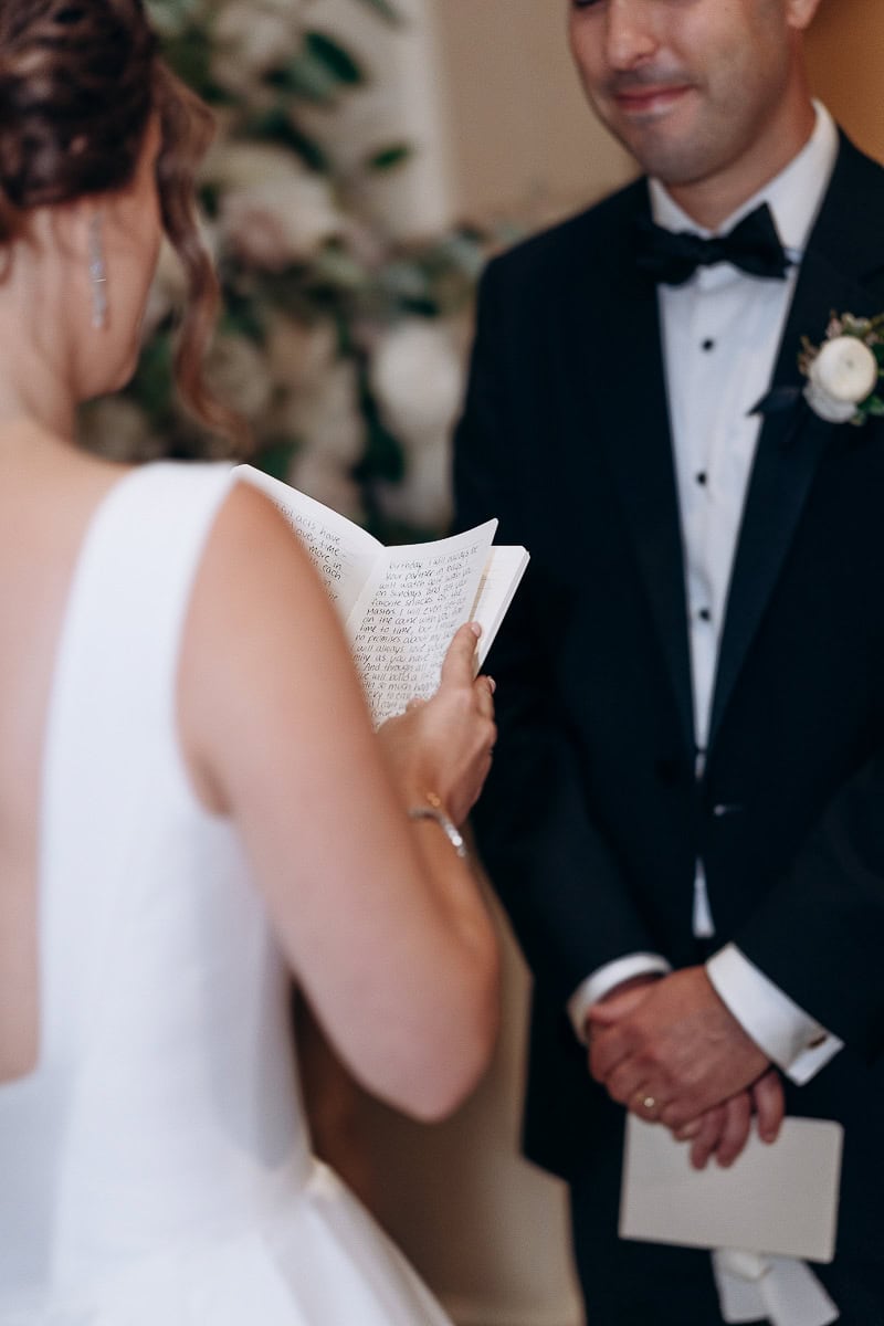 Bride in sleeveless white gown reading handwritten vows from a notebook while groom in black tuxedo listens attentively at Westchester Country Club, Rye, NY.