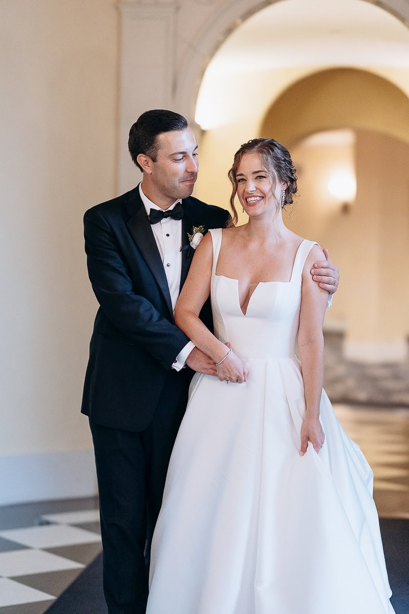 Groom in black tuxedo gently holding bride in white structured gown, standing in elegant arched hallway at Westchester Country Club, Rye, NY.