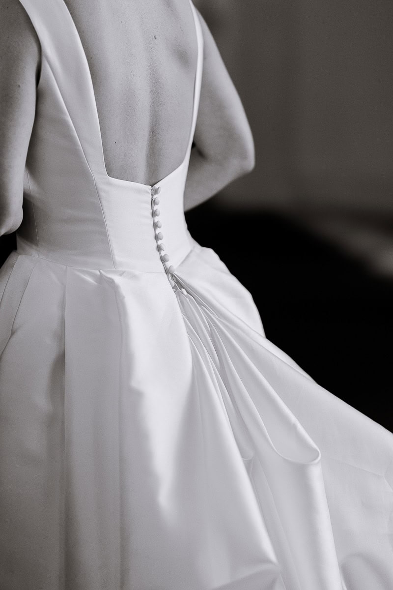 Close-up of a bride's back showing satin wedding dress with covered buttons and pleated skirt detail in soft black and white light.