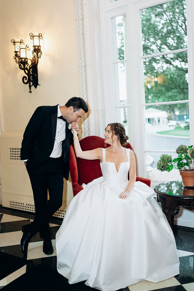 Groom in black tuxedo kissing bride's hand as she sits in red chair wearing white wedding gown near window at Westchester Country Club, Rye, NY.