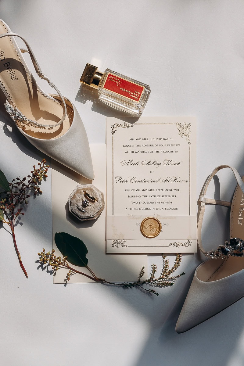 Flat lay of wedding invitation with gold wax seal, Bella Belle crystal heels, velvet ring box with rings, Maison Francis Kurkdjian perfume, and sprigs of greenery on white surface.