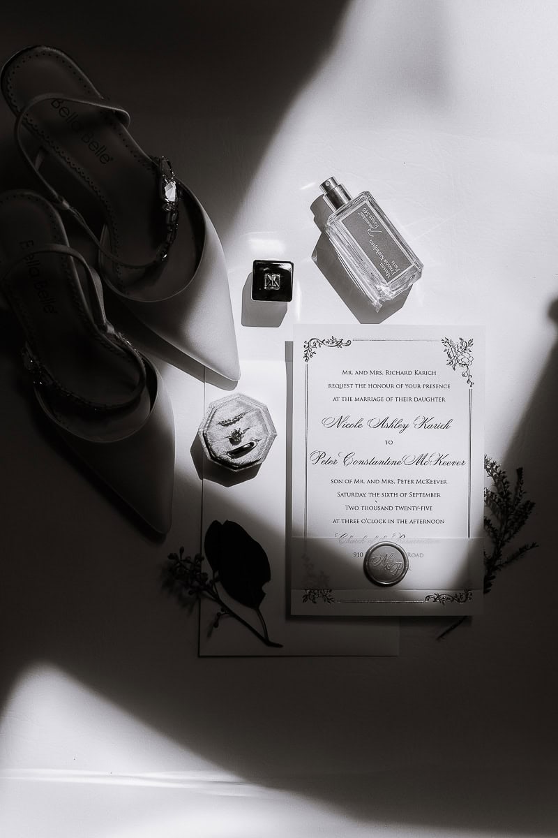 Flat lay of pointed satin bridal heels, perfume bottle, velvet ring box with rings, and an ornate wedding invitation on white surface with shadows, Westchester Country Club, Rye NY.