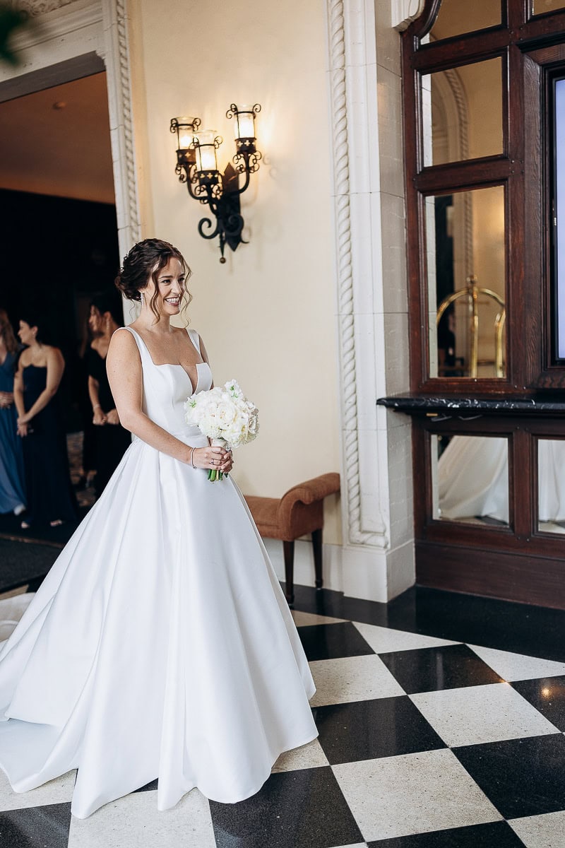 Bride in a structured white satin wedding gown holding a white flower bouquet, standing on black and white checkered floor near ornate wall sconces at Westchester Country Club, Rye, NY.