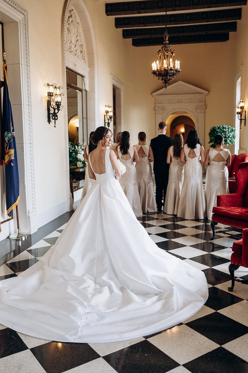 Bride in a white satin gown with long train and open back stands in a black-and-white checkered corridor with bridesmaids and groom facing away at Westchester Country Club, Rye, NY.