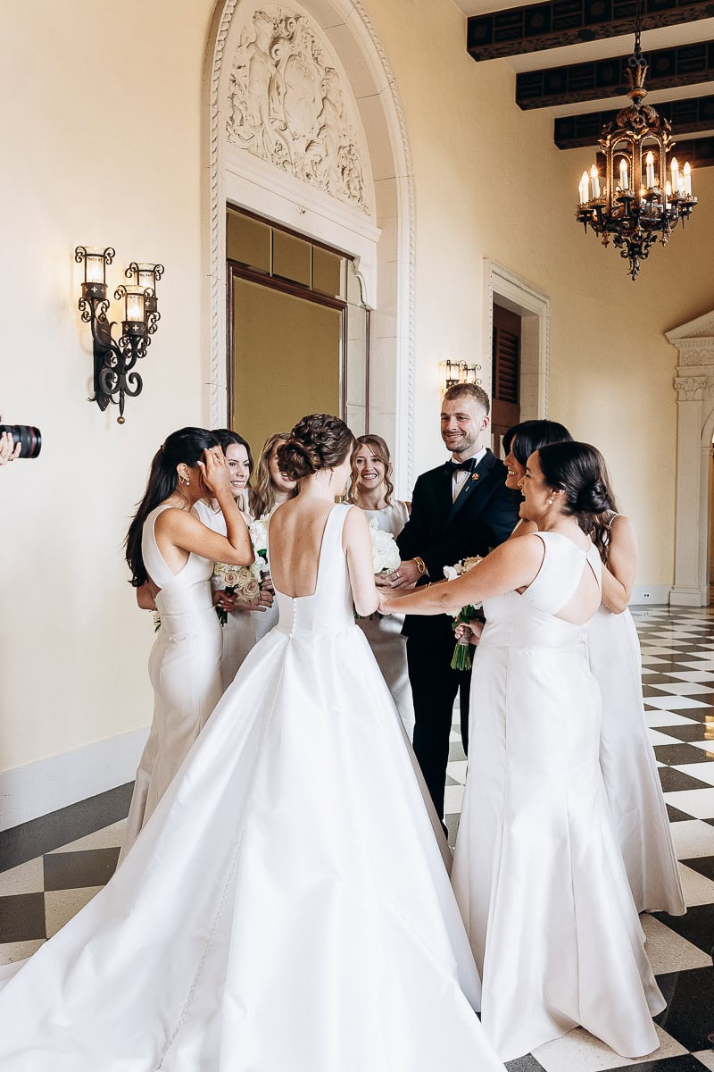 Bride in a satin gown surrounded by bridesmaids and a groomsman in a grand hall with ornate wall sconces and chandelier at Westchester Country Club, Rye, NY.
