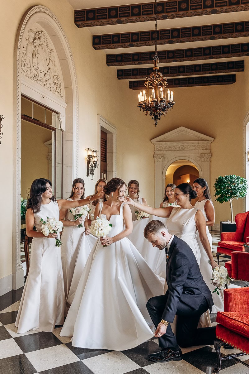 Bride in structured white gown with bridesmaids in sleek dresses and a man adjusting her dress in an elegant hallway with checkered floor and vintage chandelier at Westchester Country Club, Rye, NY.