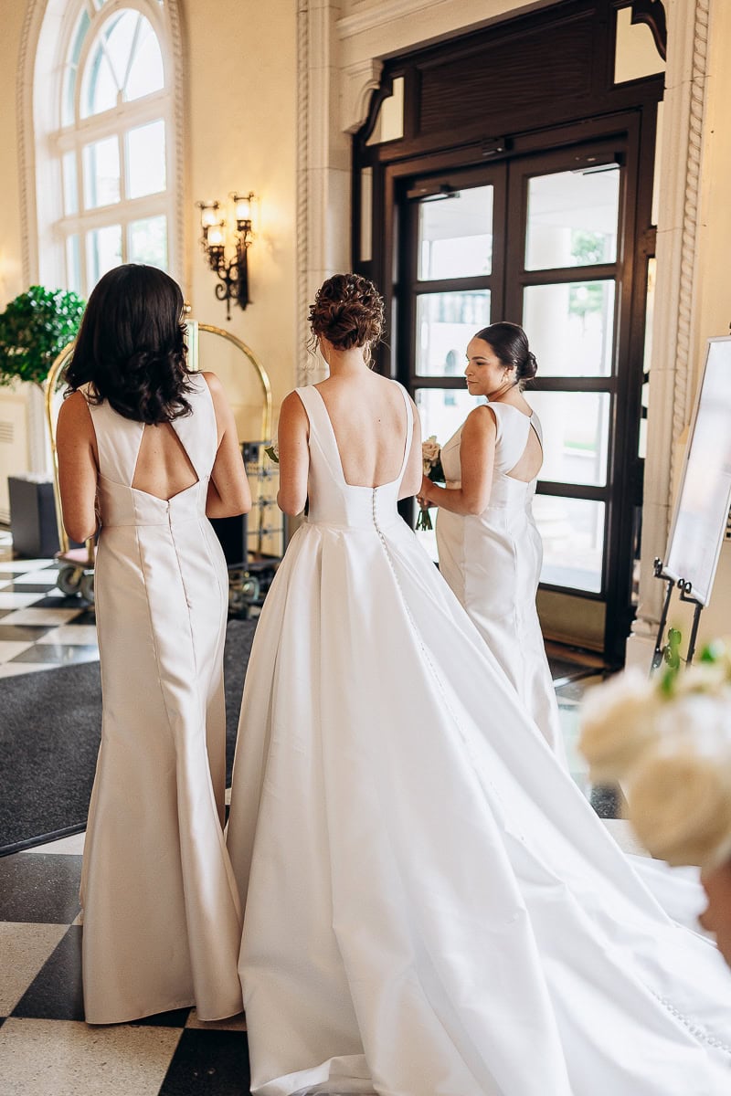 Three women in satin gowns with open backs standing inside an elegant venue with arched windows and dark wood doors at Westchester Country Club, Rye, NY.