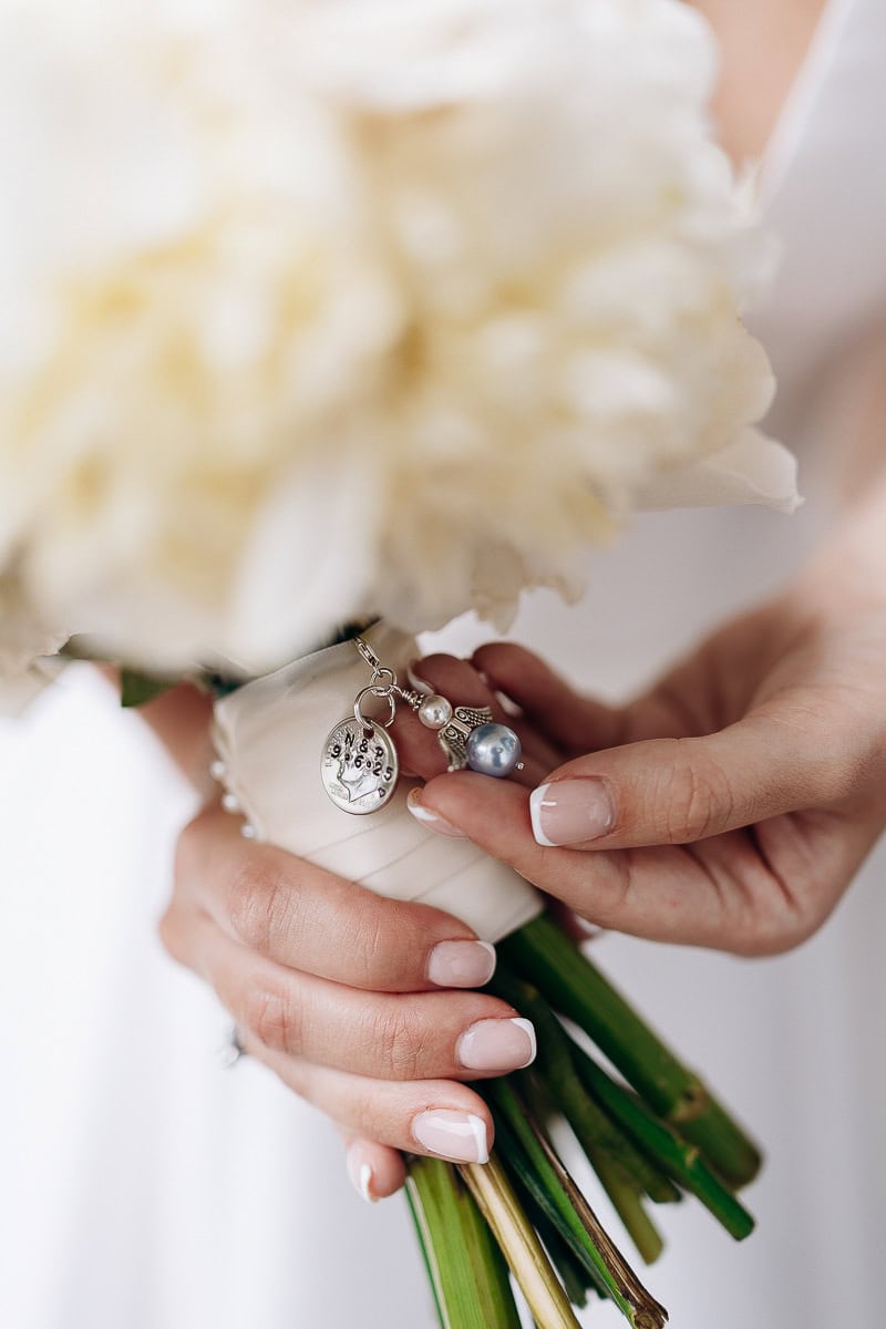 Close-up of hands holding a bridal bouquet wrapped in ivory ribbon with silver and pearl charms attached, highlighting fine French manicure and floral stems.