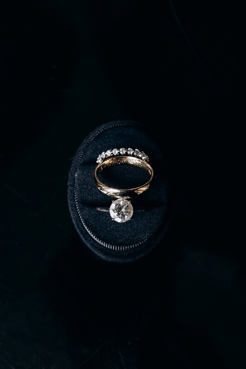 Close-up of gold wedding band, diamond eternity ring, and solitaire engagement ring on black velvet cushion.