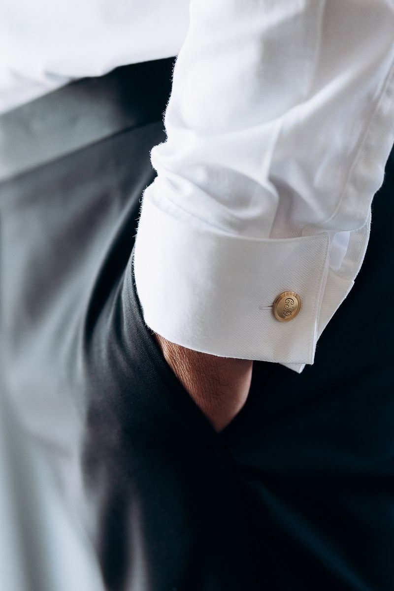 Close-up of a man's wrist with a white shirt cuff secured by a gold monogrammed cufflink, hand casually tucked into black trousers pocket.