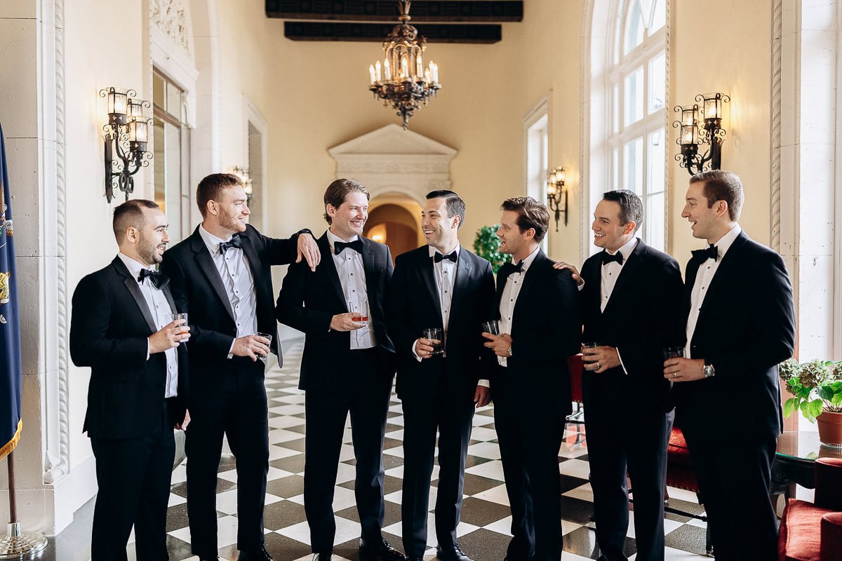 Seven men in black tuxedos holding drinks, standing in a sunlit hallway with checkered marble floor and ornate wall sconces at Westchester Country Club, Rye, NY.