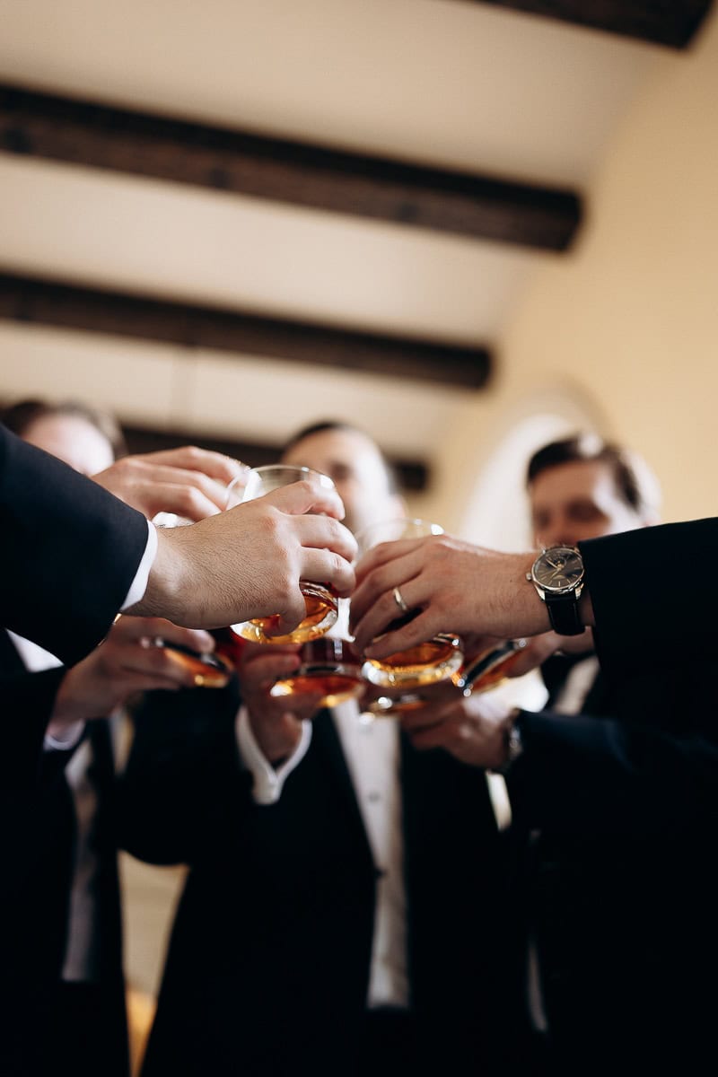 Close-up of groomsmen's hands holding whiskey glasses in a toast, with blurred faces and wooden ceiling beams in background.
