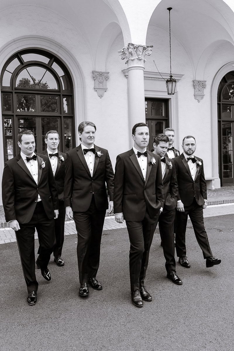 Seven men in tailored black tuxedos walking outside Westchester Country Club with white stucco walls and arched windows in the background.