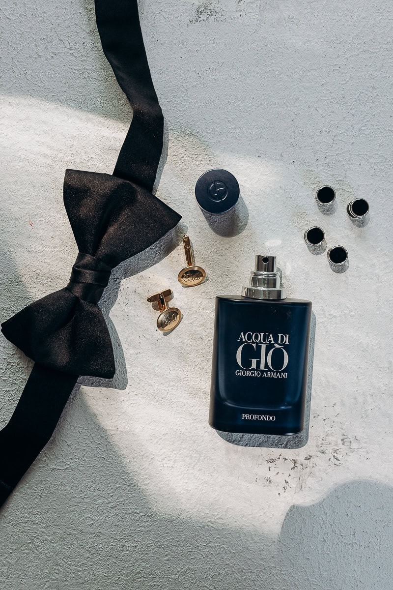 Flat lay of black silk bow tie, gold cufflinks, and Acqua di Giò Profondo cologne bottle on textured white surface