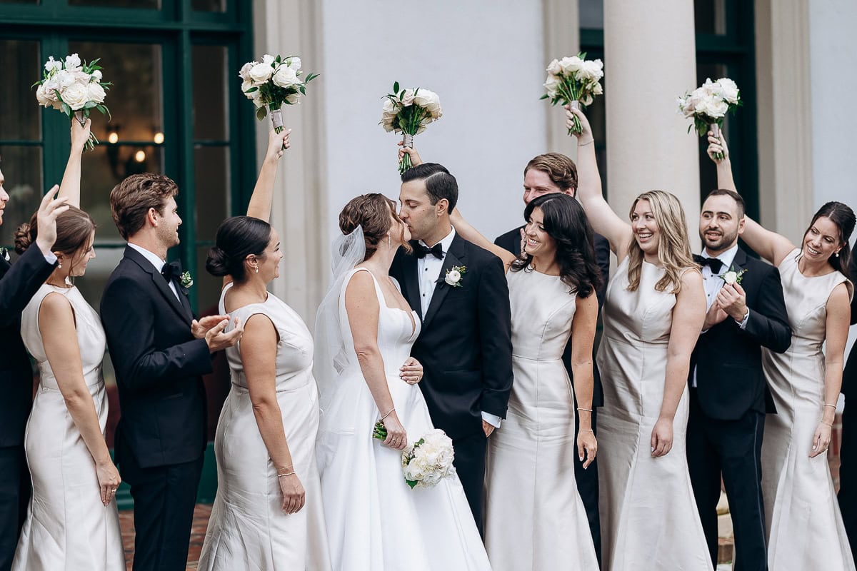 Bride and groom kissing surrounded by bridesmaids in ivory dresses and groomsmen in black tuxedos holding white flower bouquets at Westchester Country Club, Rye, NY.