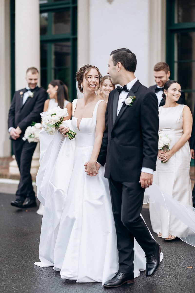 Bride in structured white gown holding bouquet, smiling at groom in black tuxedo, with bridal party in formal attire behind them at Westchester Country Club, Rye, NY.