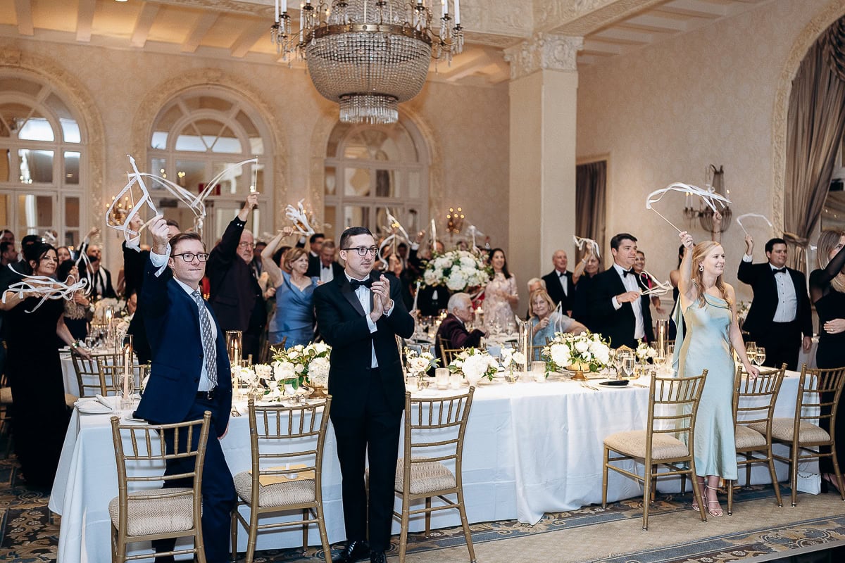Wedding guests in formal attire waving ribbon sticks at a banquet table adorned with white floral centerpieces inside an elegant ballroom at Westchester Country Club, Rye, NY.