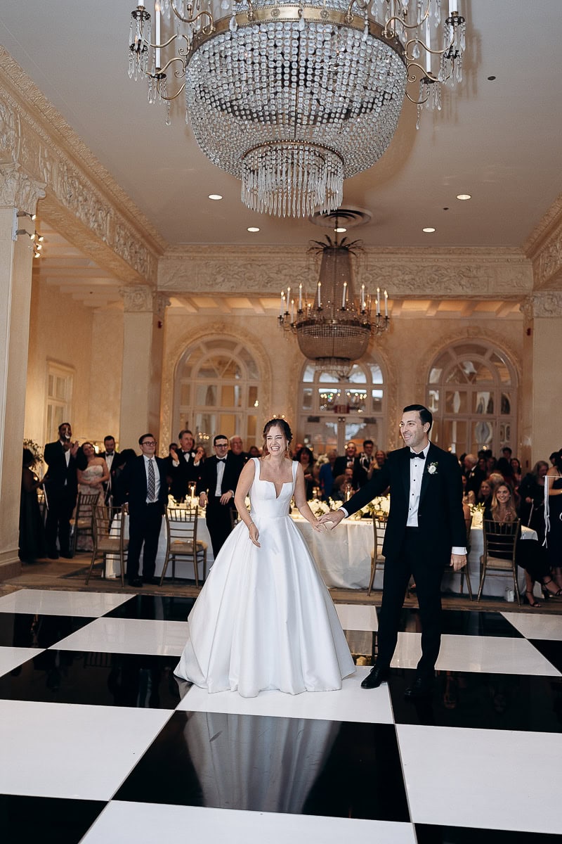 Bride in a structured white gown and groom in a black tuxedo holding hands on a black-and-white checkered dance floor with guests applauding in the background at Westchester Country Club, Rye, NY.