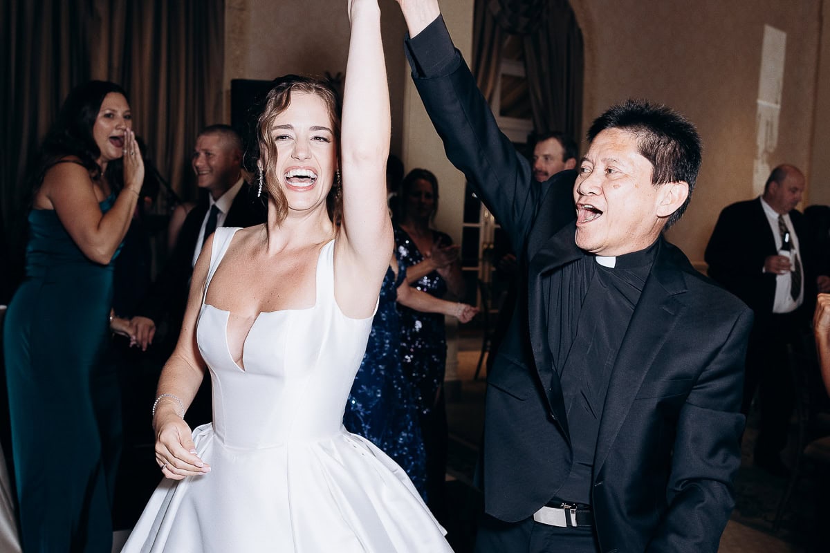 Bride in white satin wedding dress dancing with clergyman in black suit, both smiling and raising hands at Westchester Country Club reception.