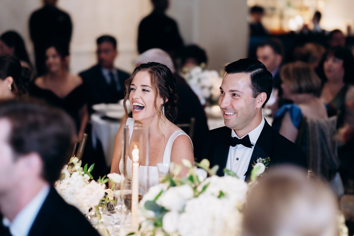 Bride and groom seated at a wedding reception table surrounded by white flowers and a lit candle, guests blurred in the background at Westchester Country Club.