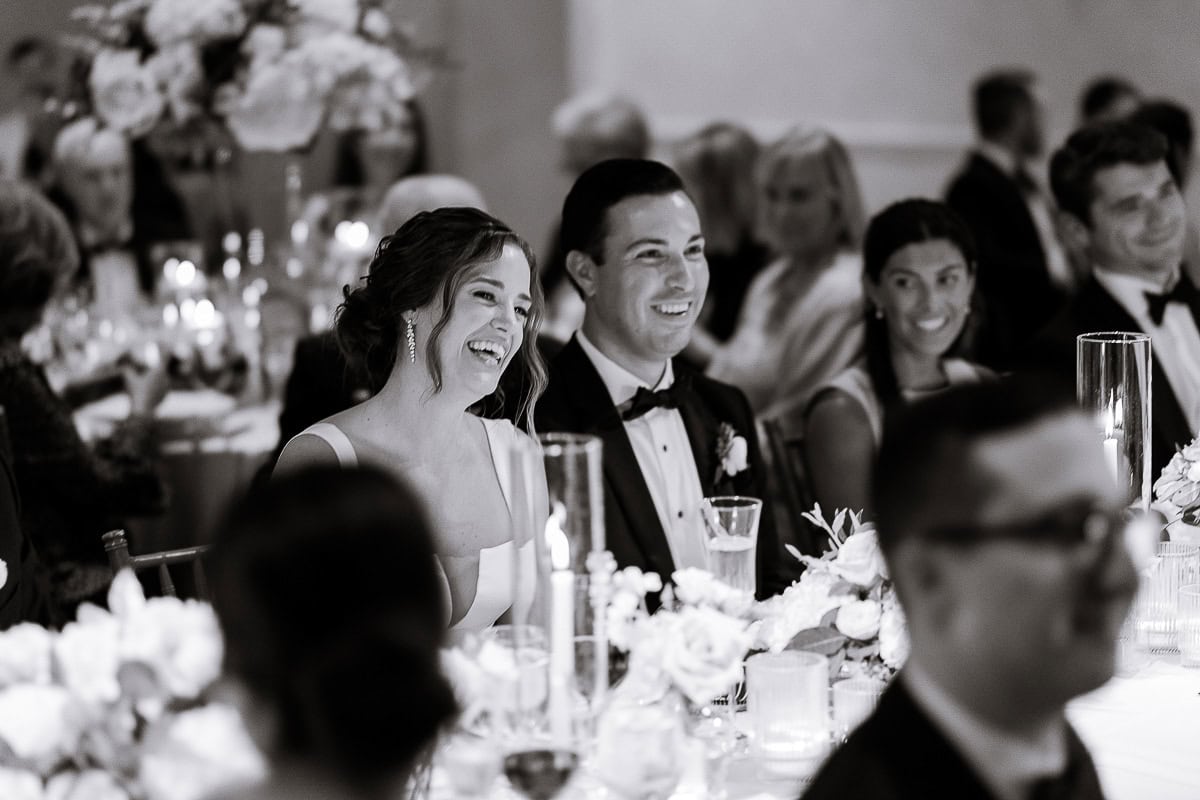 Black and white photo of a bride and groom smiling and laughing at a wedding reception table adorned with floral arrangements and candles at Westchester Country Club, Rye, NY.