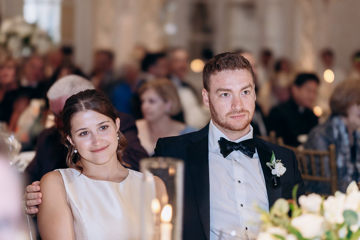 Couple seated closely at a wedding reception, the groom in a black tuxedo with a boutonniere, the bride in a sleeveless ivory dress, softly lit by candles, with blurred guests in the background at Westchester Country Club, Rye, NY.