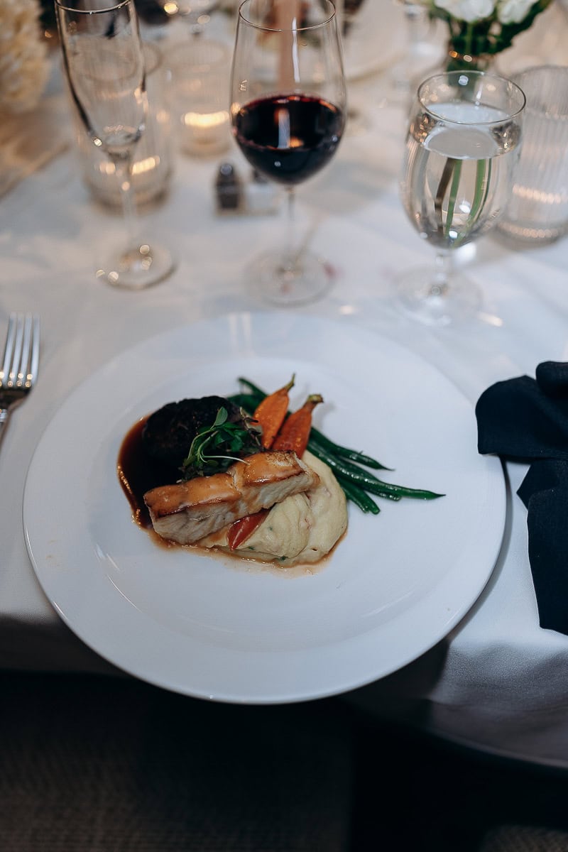 Plated gourmet entrée with seared fish, mashed potatoes, glazed carrots, green beans, and red wine glass on white tablecloth at Westchester Country Club.