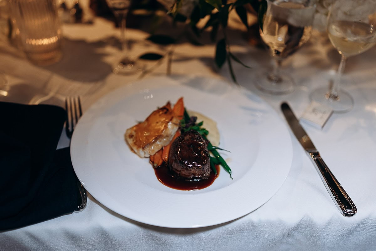 Close-up of a plated gourmet dinner with filet mignon, glazed carrots, green beans, and puree on a white plate with silverware and glassware on a white tablecloth at a formal dining setting.