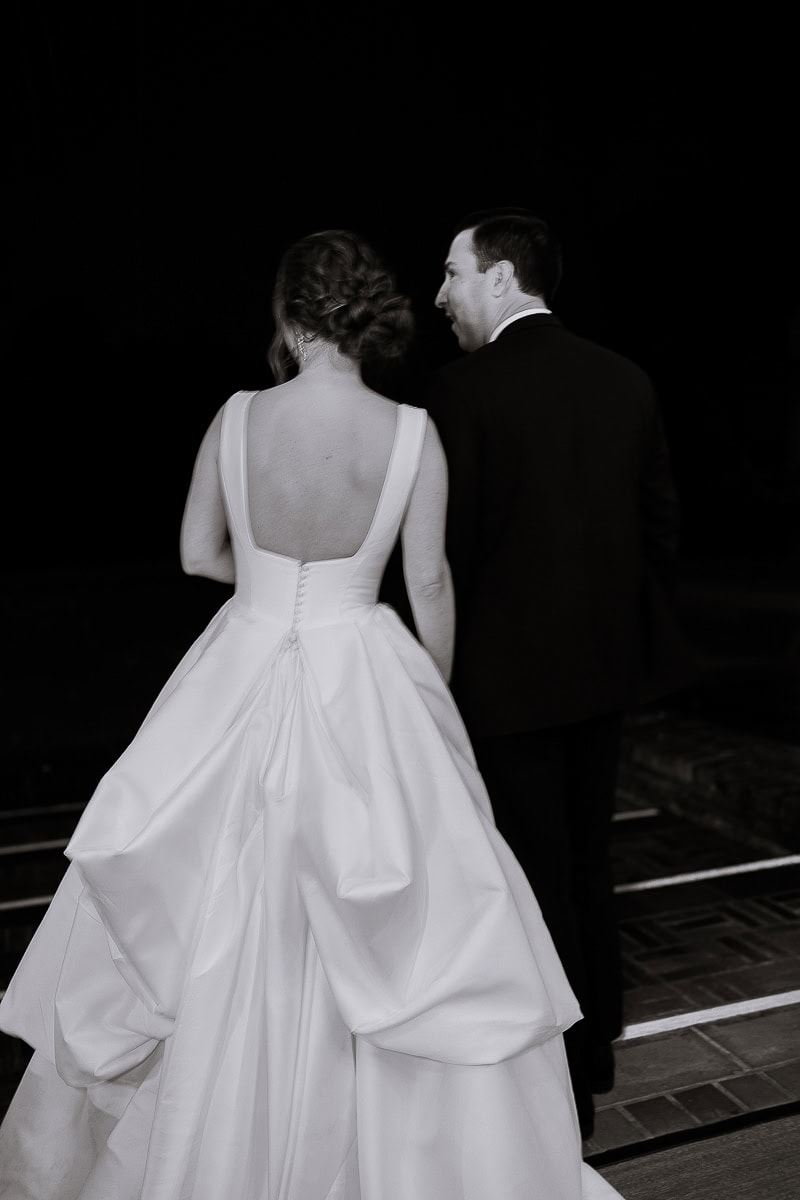 Bride in a white gown with sculptural folds and low back standing beside groom in black tuxedo at night, Westchester Country Club, Rye, NY.