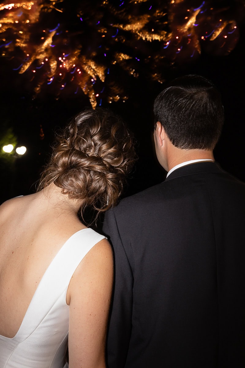 Back view of a couple in formal attire watching fireworks at night, highlighting the bride's intricate updo and the groom's tailored suit at Westchester Country Club.