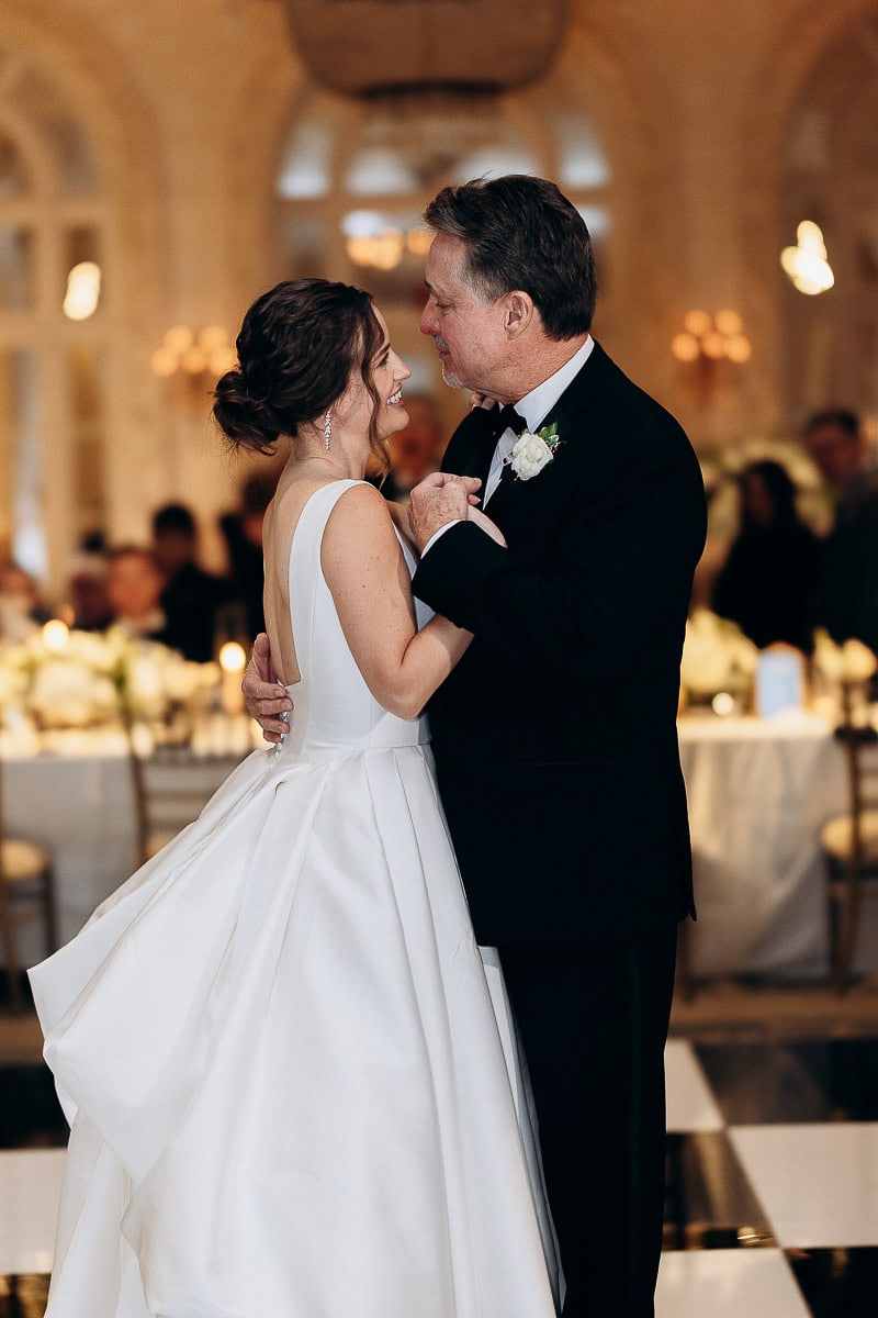 Bride in a white gown sharing an intimate dance with her father in a black tuxedo at a formal wedding reception.