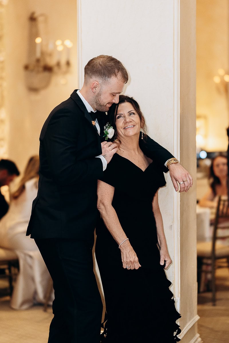 Groom in black tuxedo leaning affectionately against a woman in a black evening gown by a cream-colored pillar at Westchester Country Club, Rye, NY.