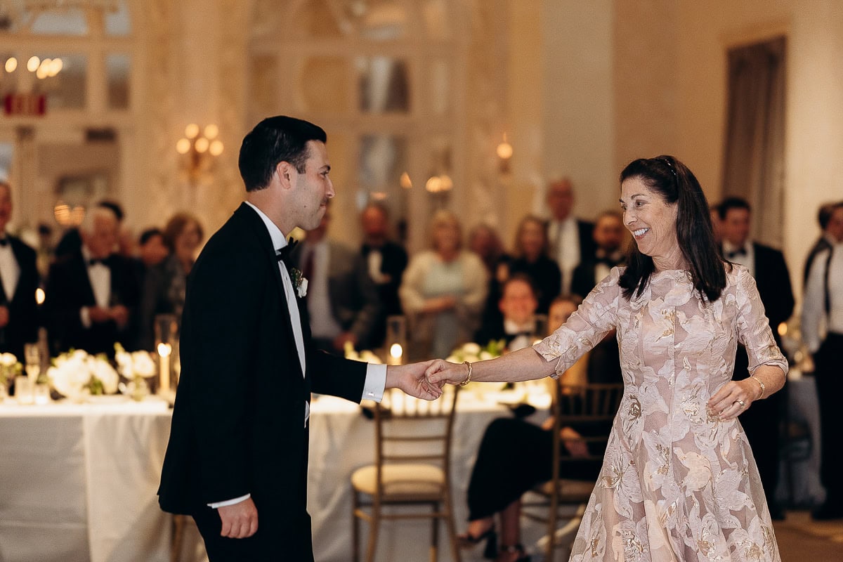 Groom in black tuxedo holding hands and dancing with his mother in a floral embroidered dress at an indoor wedding reception with guests seated and standing in the background.