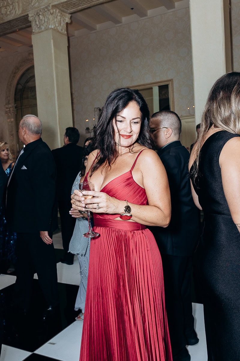 Woman in pleated red gown holding champagne glass at an indoor formal event with ornate columns and guests in the background
