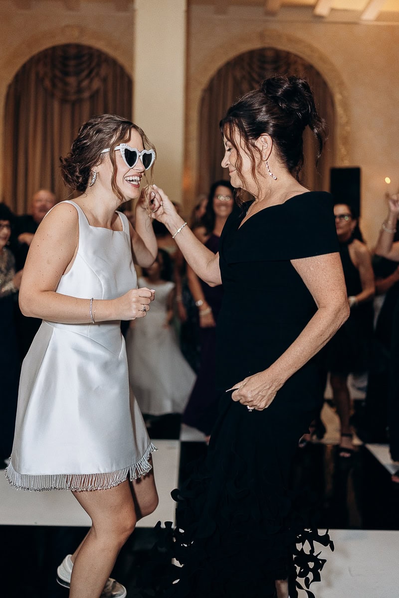 Bride wearing heart-shaped sunglasses and a white satin dress dancing with a woman in a black gown at Westchester Country Club, Rye, NY.