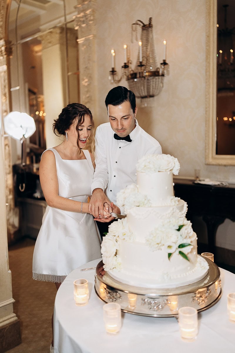 Bride and groom cutting a white three-tiered wedding cake adorned with white flowers, surrounded by candlelight in an elegant room at Westchester Country Club.