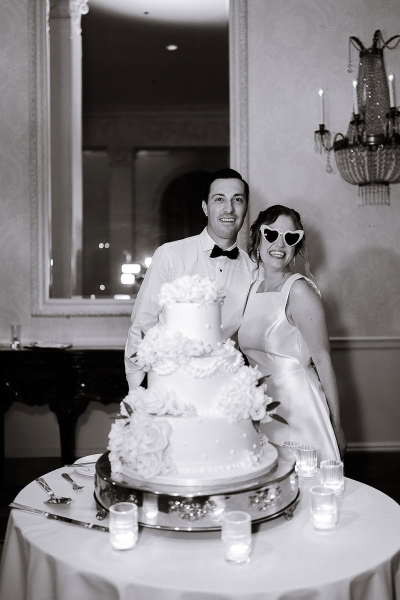Bride in satin gown with heart-shaped sunglasses and groom in tuxedo standing behind a three-tier floral wedding cake on a round table with candles at Westchester Country Club.