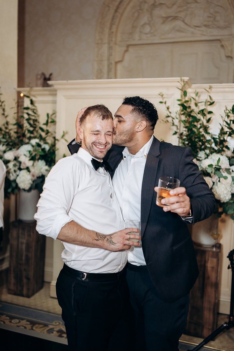 Two men in formal attire share a tender moment; one kisses the other on the temple while holding drinks, surrounded by floral decor at Westchester Country Club.