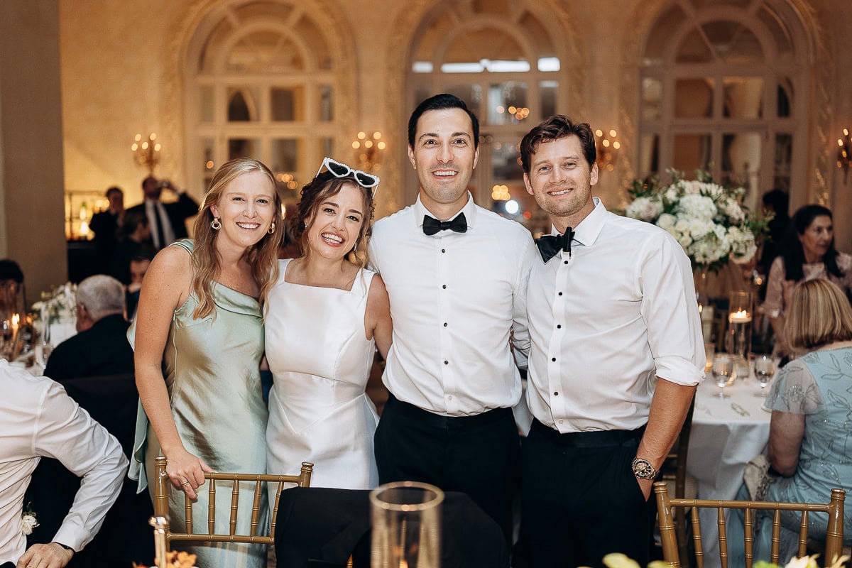 Four guests in formal attire smiling at a wedding reception inside Westchester Country Club, Rye, NY, with soft lighting and floral arrangements in the background.